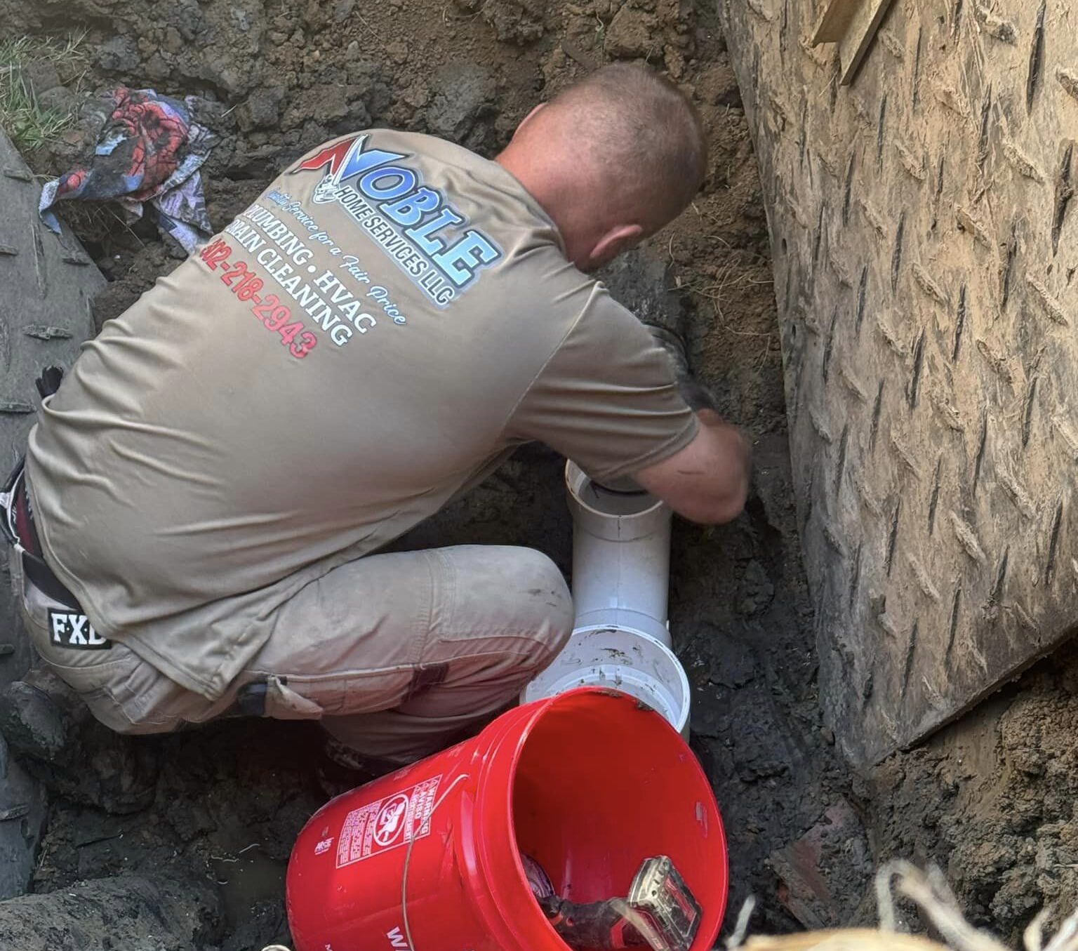 Plumber from Noble Home Services LLC working on a pipe installation in a trench, with a red bucket nearby, emphasizing skilled plumbing services in Townsend, DE.
